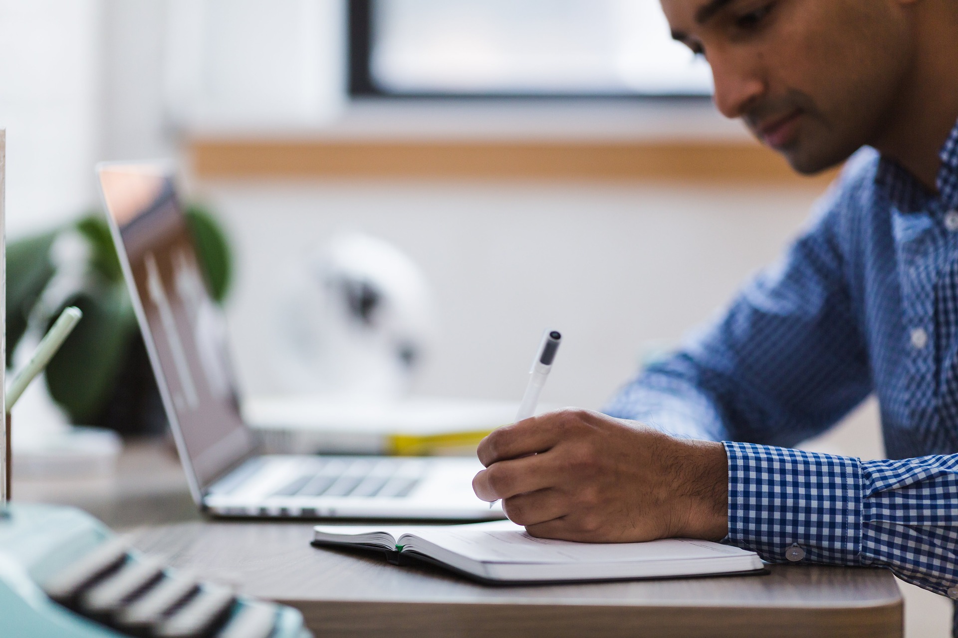 Male sat at a desk with a laptop blurred in the backround, writing in a notebook, intended to reflect a lawyer making notes and taking action after coaching for solicitors and other fee earners.